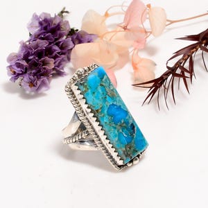 May include: A silver ring with a large, rectangular turquoise stone. The stone is a vibrant blue with darker matrix patterns. The ring has a detailed, textured silver setting with a rope-like border and a decorative design around the stone.