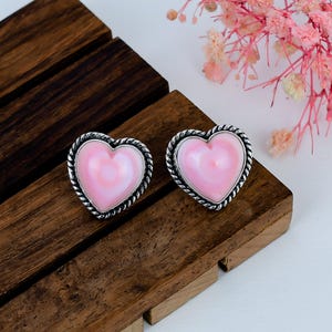 May include: A pair of heart-shaped stud earrings featuring smooth, light pink centers set within a textured, twisted silver-toned metal border. The earrings have a polished finish and a distinct rope-like edge surrounding the pink heart inlay.