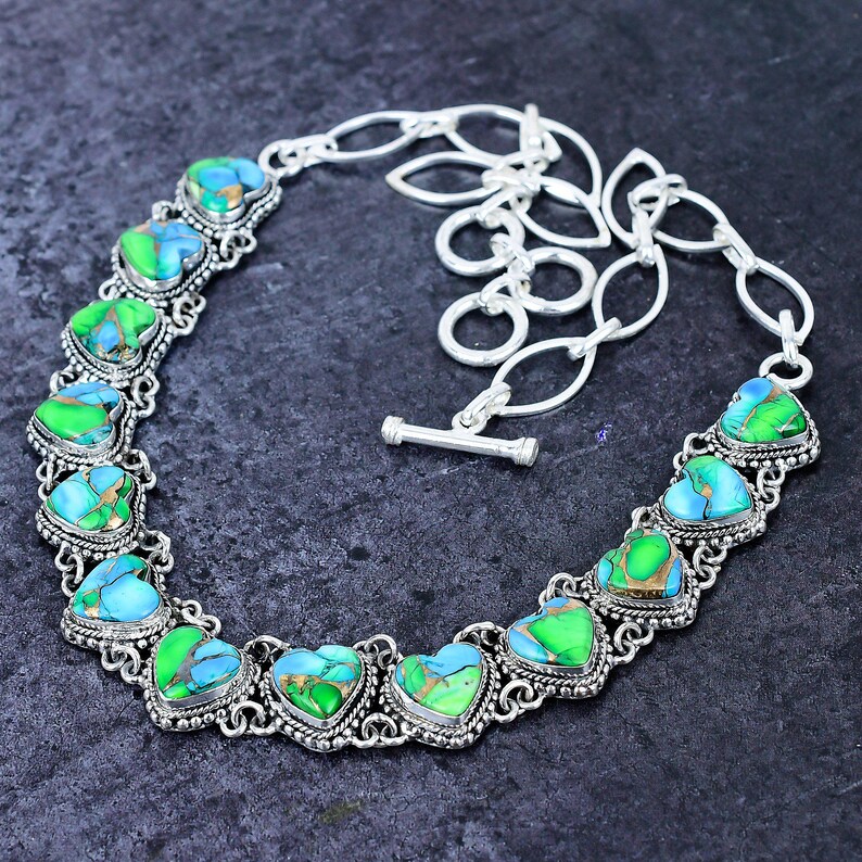 May include: A silver-toned necklace featuring heart-shaped turquoise and green stones. The stones are set in ornate, filigree-style settings. The necklace has a toggle clasp and a chain-link extension.