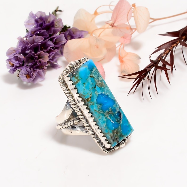 May include: A silver ring with a large, rectangular turquoise stone. The stone is a vibrant blue with darker matrix patterns. The ring has a detailed, textured silver setting with a rope-like border and a decorative design on the band.
