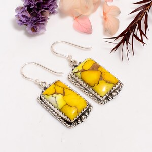 May include: Silver earrings featuring rectangular pendants with yellow and brown stones. The stones are set in a decorative silver frame with a beaded edge. The earrings have a hook closure. The image shows the earrings on a white surface with floral accents.