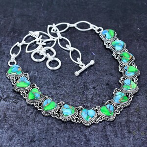 May include: A silver-toned necklace featuring heart-shaped turquoise and green stones. The stones are set in a decorative silver frame and connected by a chain. The necklace has a toggle clasp.