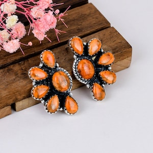 May include: A pair of stud earrings featuring clusters of orange, oval-shaped stones set in oxidized silver. Each earring has a central oval stone surrounded by smaller, teardrop-shaped stones with a scalloped, rope-textured silver border.