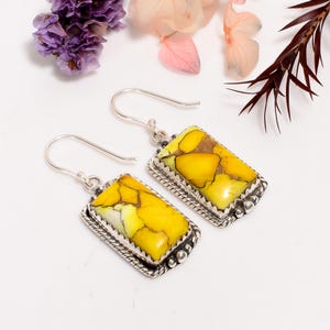 May include: Silver earrings featuring rectangular pendants with yellow and brown stones. The stones are set in a decorative silver frame with a beaded edge. The earrings have a hook closure. The image shows the earrings on a white surface with floral accents.