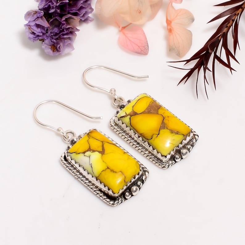 May include: Silver earrings featuring rectangular pendants with yellow and brown stones. The stones are set in a decorative silver frame with a beaded edge. The earrings have a hook closure. The image shows the earrings on a white surface with floral accents.