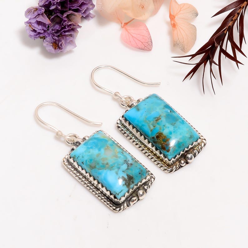 May include: A pair of turquoise and silver earrings. Each earring features a rectangular turquoise stone with brown veining, set in a silver frame with a decorative edge. The earrings have a hook closure.