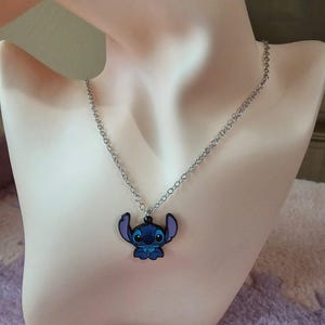 May include: A silver-colored chain necklace with a blue and black Stitch pendant. The pendant features the cartoon character Stitch from the Disney movie Lilo & Stitch. The necklace is displayed on a mannequin.