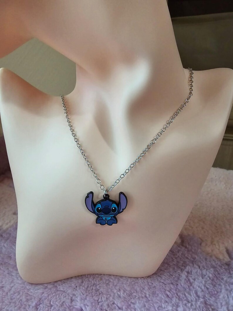 May include: A silver-colored chain necklace with a blue and black Stitch pendant. The pendant features the cartoon character Stitch from the Disney movie Lilo & Stitch. The necklace is displayed on a mannequin.