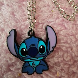 May include: A blue and black Stitch pendant necklace with a silver chain. The pendant features the character Stitch from the Disney movie Lilo & Stitch, with blue, black, and purple accents. The necklace is displayed on a pink, fuzzy surface.