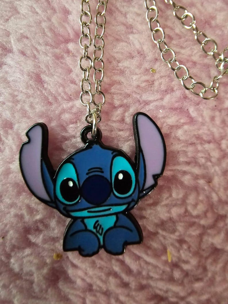 May include: A blue and black Stitch pendant necklace with a silver chain. The pendant features the character Stitch from the Disney movie Lilo & Stitch, with blue, black, and purple accents. The necklace is displayed on a pink, fuzzy surface.