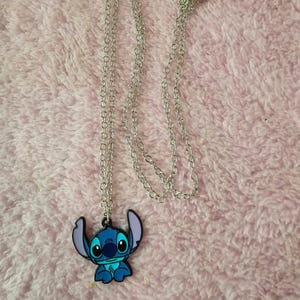 May include: A silver-colored chain necklace with a blue and black Stitch pendant. The pendant features the cartoon character Stitch with large eyes and ears. The necklace is displayed on a pink, fuzzy surface.