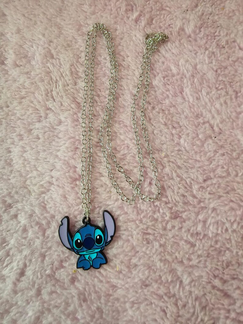 May include: A silver-colored chain necklace with a blue and black Stitch pendant. The pendant features the cartoon character Stitch with large eyes and ears. The necklace is displayed on a pink, fuzzy surface.