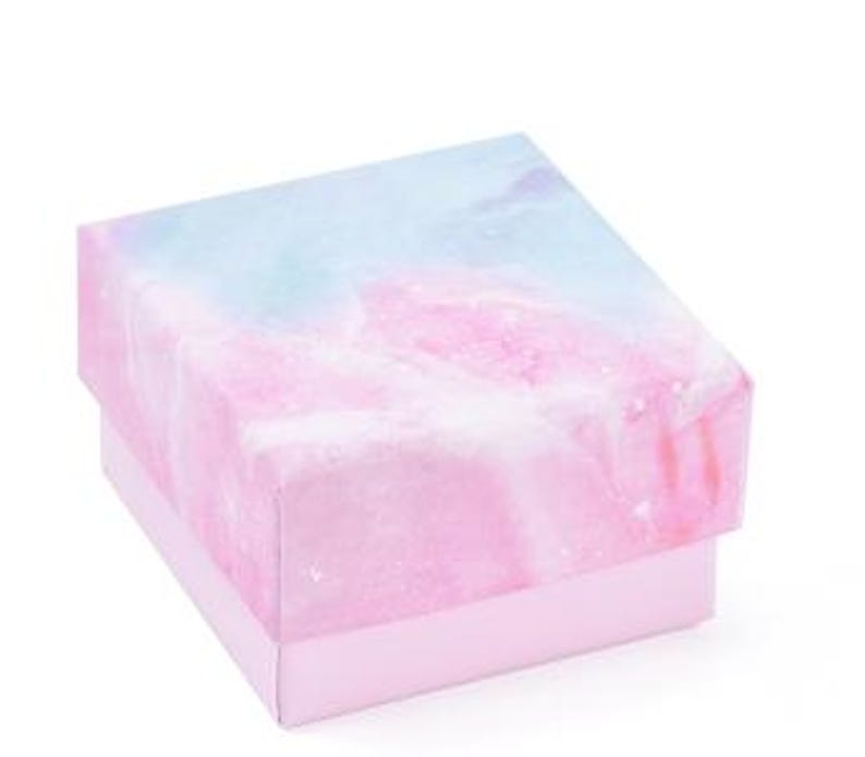 May include: A square gift box with a pastel watercolor design. The lid features a blend of pink, blue, and white hues, resembling a marble or galaxy pattern. The box's base is a solid light pink color.