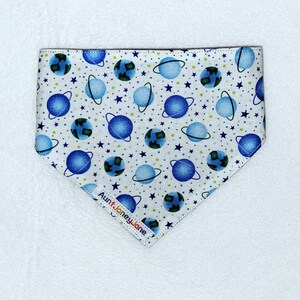 May include: White bandana with a blue and green space-themed print. The bandana features planets, stars, and rings. The bandana has a white label with the text "Aunt Janey Jane" in rainbow colors.