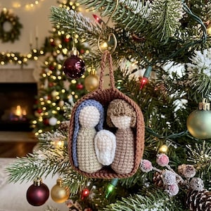 May include: A handmade crochet Christmas ornament depicting the Nativity scene. The ornament features three figures: Mary, Joseph, and baby Jesus, all enclosed in a brown woven basket. The ornament is hanging on a Christmas tree with other ornaments and lights.
