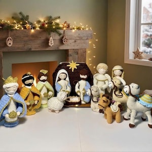 May include: A crochet nativity set featuring figures of Mary, Joseph, the baby Jesus in a manger, three wise figures, an angel, a sheep, and two camels. The figures are crafted with yarn in various colors including blue, yellow, brown, and white, arranged in a group with a dark brown crochet stable structure.