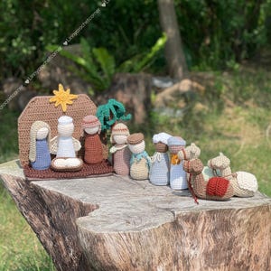 Può includere: Un presepe all'uncinetto con personaggi in vari colori, tra cui blu, marrone e bianco. La scena comprende una stalla, una stella e cammelli. Le figure sono disposte su una base marrone, posta su un ceppo d'albero.