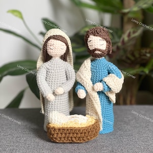 Könnte beinhalten: Gehäkelte Krippenszene mit Maria in einem grauen Gewand und Josef in einem blauen Gewand mit einem cremefarbenen Schal. Ein Baby in einer braunen Wiege ist vor ihnen. Die Figuren haben geschlossene Augen. Im Hintergrund eine grüne Pflanze.