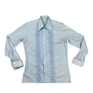 Pode incluir: Camisa azul clara de manga comprida com gola pontuda e punhos abotoados. Apresenta um design bordado vertical em um tom de azul mais escuro na frente. A camisa é feita de tecido leve.