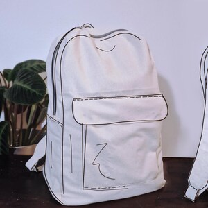 Może przedstawiać: Dwa białe plecaki z czarnymi obrysami. Jeden plecak jest pokazany z przodu z kieszenią i zamkiem błyskawicznym. Drugi plecak jest pokazany od tyłu z widocznymi paskami. Plecaki znajdują się na drewnianej powierzchni.