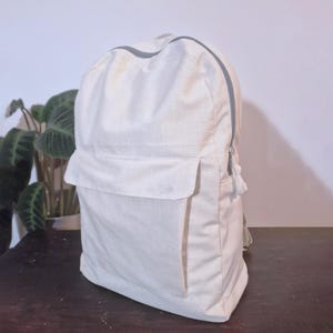 Puede incluir: Una mochila de color crema con un bolsillo frontal rectangular y una cremallera gris. La mochila tiene un dise&ntilde;o sencillo y parece estar hecha de una tela resistente. La cremallera recorre la parte superior y el lateral de la bolsa.