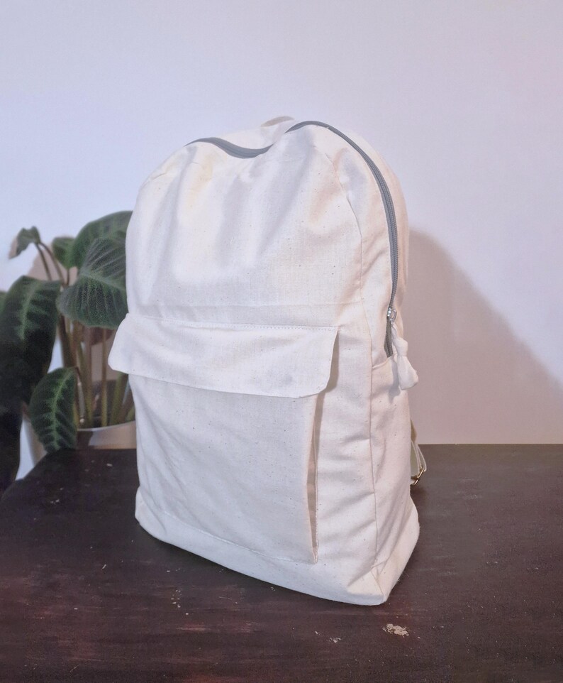 Puede incluir: Una mochila de color crema con un bolsillo frontal rectangular y una cremallera gris. La mochila tiene un dise&ntilde;o sencillo y parece estar hecha de una tela resistente. La cremallera recorre la parte superior y el lateral de la bolsa.