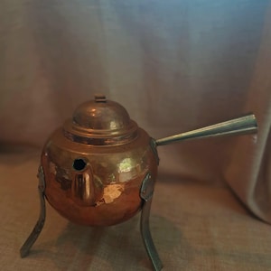 Könnte beinhalten: Eine Kupferteekanne mit rundem Körper, Deckel und langem, silberfarbenem Ausguss. Die Teekanne hat drei silberfarbene Beine und steht vor einem neutralen Hintergrund. Die Oberfläche der Teekanne hat eine gehämmerten Textur.