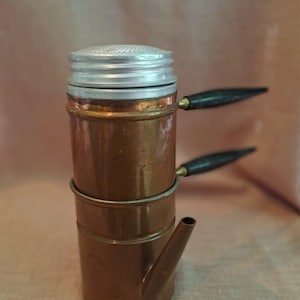 Pode incluir: Cafeteira de cobre vintage com tampa prateada e alças de madeira preta. A cafeteira tem um corpo cilíndrico com bico e um anel de metal. Um item de cozinha clássico.