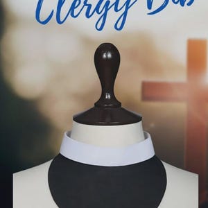 May include: A black clergy bib with a white collar, displayed on a mannequin head. The words "Clerical Collection" and "Clergy Bib" are written in blue script at the top of the image. The background is blurred with a cross.
