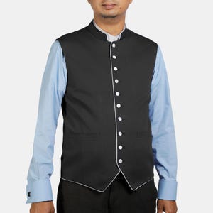May include: A black vest with white trim and buttons, worn over a light blue dress shirt. The vest has a mandarin collar and two front pockets. The person is wearing black pants.