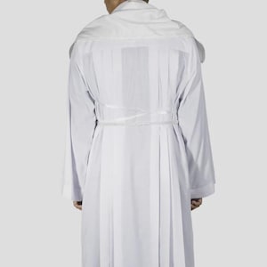 May include: A white, long-sleeved robe with a pleated skirt and a decorative belt. The robe has a high collar and is worn by a person facing away from the camera. The background is a solid, light gray.