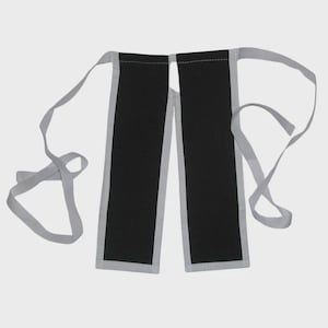 May include: A black apron-like garment with gray trim and long gray ties. The apron has two rectangular panels connected at the top. The gray trim borders the black panels. The ties extend from the top corners.