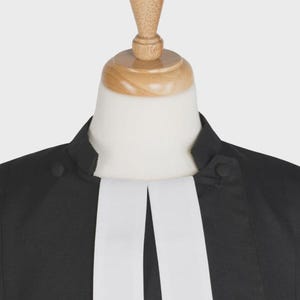 May include: Black clergy shirt with a white clerical collar and a wooden neck form. The shirt has a button closure and a simple, elegant design. The white collar is a prominent feature.