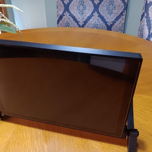 May include: A rectangular black digital display screen mounted on a black adjustable stand. The screen has a dark, reflective surface and is positioned at an angle. The frame surrounding the display is thin and black, with two sturdy black supports.