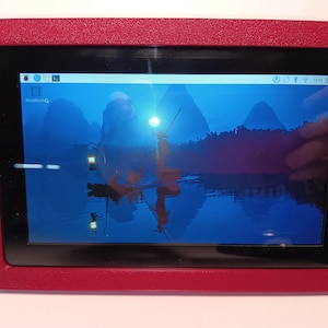 May include: A rectangular electronic display screen encased in a textured, matte red frame. The screen shows a digital desktop interface with a blue landscape wallpaper featuring a silhouette of a person on a boat, a lantern, and mountains.