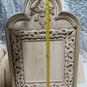 May include: A light-colored, unfinished wooden carving of a religious icon. The carving features a dove with outstretched wings at the top, and ornate scrollwork around the edges. The height is approximately 27 inches.