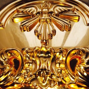 May include: Close-up of a gold-colored, ornate decorative element. The design features a central stylized sunburst motif with radiating lines, and curved, symmetrical flourishes. The surface reflects light, highlighting the intricate details and textures.