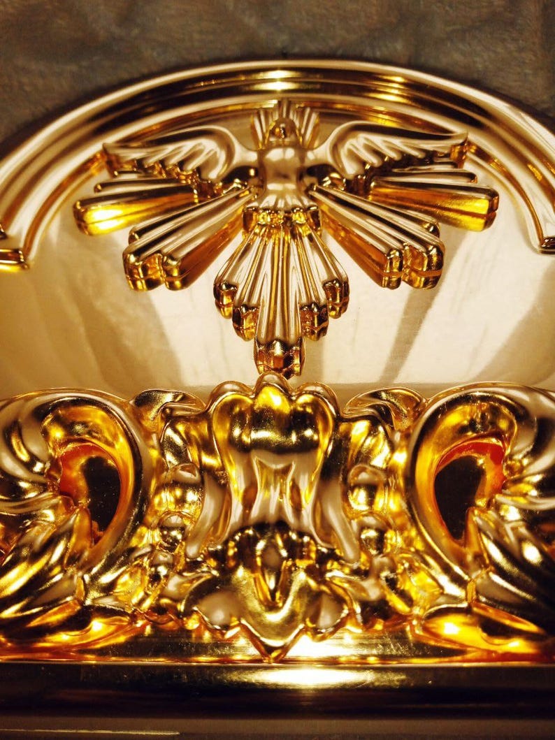 May include: Close-up of a gold-colored, ornate decorative element. The design features a central stylized sunburst motif with radiating lines, and curved, symmetrical flourishes. The surface reflects light, highlighting the intricate details and textures.