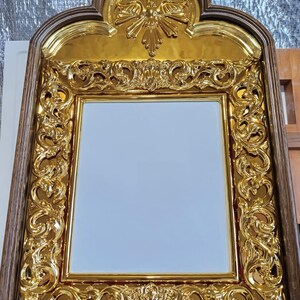 May include: Ornate gold-colored framed mirror with a rectangular mirror surface. The frame features intricate, swirling patterns and a decorative top with a dove emblem. The frame is set within a dark brown wooden border.