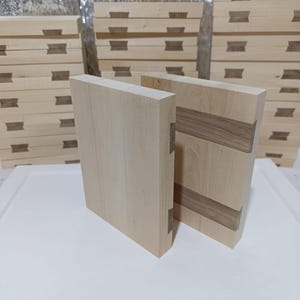 May include: Two light-coloured wooden blocks with dovetail joints are displayed, showcasing the craftsmanship. The blocks are rectangular and have a natural wood grain. Stacks of similar blocks are visible in the background, suggesting a woodworking project.