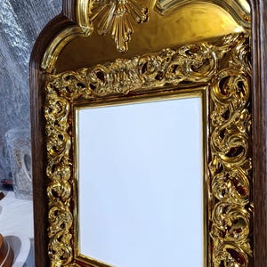 May include: Ornate, gold-colored religious icon with a dark brown wooden frame. The icon features a central white space, surrounded by intricate gold filigree and a gold dove at the top. The frame has a decorative, carved design.