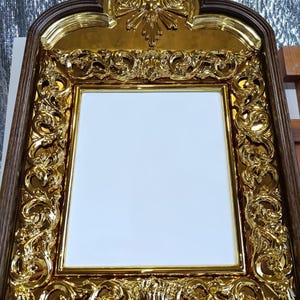 May include: Ornate gold-colored framed mirror with intricate scrollwork and a dark brown wooden border. The top of the frame features a decorative gold emblem. The mirror's surface is white.