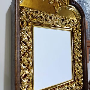 May include: Ornate gold-colored frame with a white center. The frame has intricate, swirling designs and is set within a dark brown wooden border. A gold decorative element is at the top. The frame is likely for a picture or artwork.