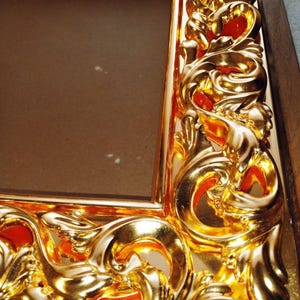 May include: Close-up of a gold-colored picture frame with ornate, swirling details. The frame has a glossy finish and a rich, warm tone. The design features intricate curves and patterns, adding a touch of elegance to the frame.