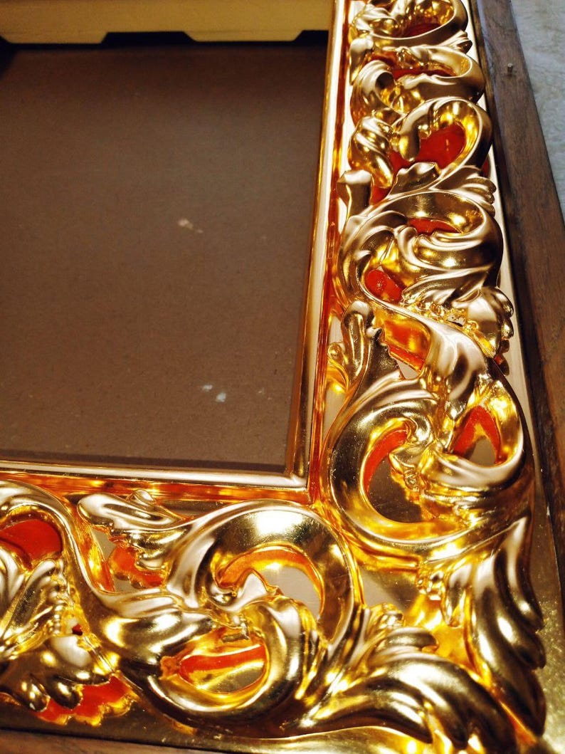 May include: Close-up of a gold-colored picture frame with ornate, swirling details. The frame has a glossy finish and a rich, warm tone. The design features intricate curves and patterns, adding a touch of elegance to the frame.