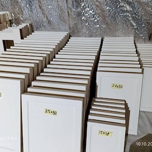 May include: A selection of white wooden picture frames in various sizes, including 15x18, 20x25, 21x30, 25x30, and 30x35 cm. Some frames have dimensions labeled with yellow stickers. The frames are stacked and ready for use.