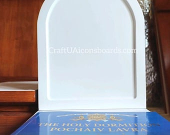 May include: A white, arched wooden icon board with a recessed panel, resting on a book with a blue cover and gold lettering. The book features an image of a church.