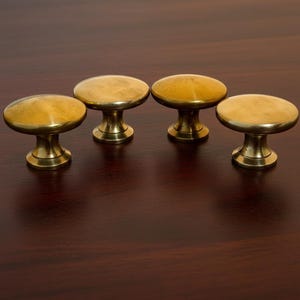 Puede incluir: Cuatro tiradores de armario de latón con una parte superior redondeada y una base corta en forma de pedestal. Los tiradores son de color dorado cálido y están dispuestos en fila. Probablemente son para muebles o armarios.