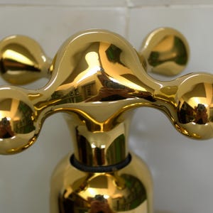 Puede incluir: Primer plano de un tirador de grifo dorado y brillante. El tirador tiene un diseño único con cuatro extremos redondeados y bulbosos. La superficie refleja el entorno, creando un efecto de espejo. El grifo parece ser de metal.