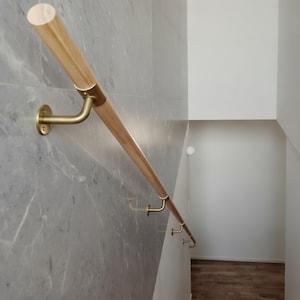 Op de afbeelding: Een houten leuning met messingkleurige metalen steunen is bevestigd aan een grijze muur met marmerpatroon. De leuning is lichtbruin en cilindrisch, met een gladde afwerking. De messing steunen zijn gebogen en aan de muur bevestigd.